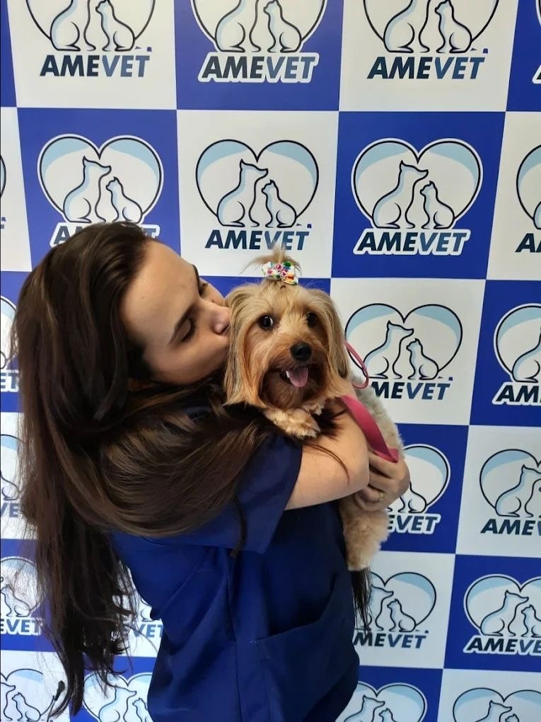 Veterinária da AMEVET abraçando um cachorro pequeno em frente ao painel da clínica